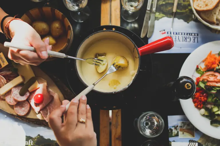 Ein Fonduetopf auf einem Tisch. Zwei Hände tauchen mit Fonduegabeln Kartoffeln im Topf. 